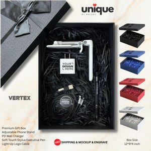 Puede incluir: Una caja de regalo negra que contiene un soporte para teléfono blanco ajustable, un cargador de pared blanco con el texto YOUR DESIGN HERE, un cable de carga negro con una etiqueta circular y un bolígrafo negro. La caja mide 30,48 x 22,86 x 10,16 cm.