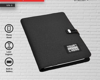 May include: A black rectangular notepad with a textured fabric cover, featuring a black strap closure with a white accent. A small black rectangular patch on the cover displays the white text YOUR DESIGN HERE. The notepad includes a built-in battery.
