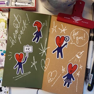 Könnte beinhalten: Notizbücher mit herzförmigen Charakteraufklebern. Eines ist olivgrün mit den Worten "heart pack!" und einem Charakter, der ein "ok"-Schild hält. Das andere ist beige mit den Worten "for you!"