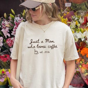 May include: An ivory short-sleeve t-shirt with black text that reads Just a Mom who loves coffee est. 2026, featuring a small coffee cup graphic. The shirt is paired with a beige and brown baseball cap, black shorts, white socks, and white trainers.