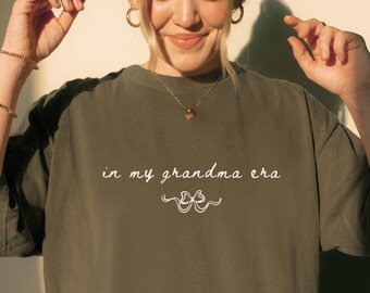 May include: A pepper-coloured t-shirt featuring the text "in my grandma era" in a script font. The shirt has a relaxed fit and short sleeves. The text is above a decorative flourish.