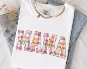 May include: A white short-sleeved t-shirt featuring the word MAMA in a multicoloured plaid pattern with a stitched border. The shirt is layered over a pair of light blue denim jeans, and a thin brown cord necklace rests on the collar area.