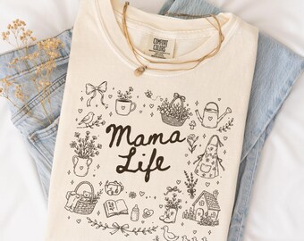 May include: A cream-coloured t-shirt featuring the text "Mama Life" in black, with hand-drawn illustrations of gardening items and a house. The t-shirt is folded on blue jeans, with a gold necklace and dried flowers. The image is set on a white sheet.