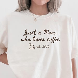 May include: Cream-colored t-shirt with the text "Just a Mom who loves coffee" and a coffee cup graphic. The text "est. 2026" is below the graphic. The person is wearing red and white checkered shorts.