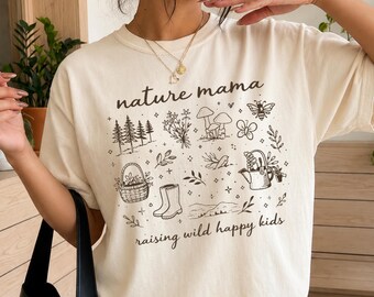 May include: An ivory short-sleeved t-shirt featuring a black graphic print with the text nature mama and raising wild happy kids. The design includes illustrations of pine trees, wildflowers, mushrooms, a bee, a basket, boots, and a watering can.