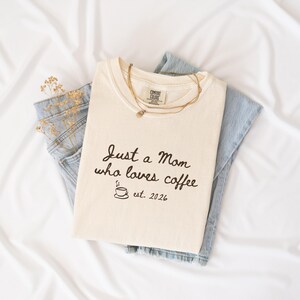 May include: A cream-coloured t-shirt featuring the black text Just a Mom who loves coffee, a small coffee cup icon, and the text est. 2026. The shirt is layered over light blue denim jeans and accompanied by a thin gold-toned layered necklace.