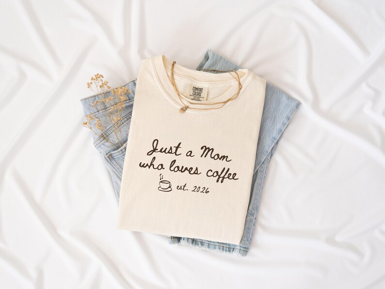 May include: A cream-coloured t-shirt featuring the black text Just a Mom who loves coffee, a small coffee cup icon, and the text est. 2026. The shirt is layered over light blue denim jeans and accompanied by a thin gold-toned layered necklace.