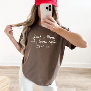 May include: A brown short-sleeved t-shirt featuring white cursive text that reads Just a Mom who loves coffee, accompanied by a small line-art coffee cup icon and the text est. 2026, paired with a bright red baseball cap.