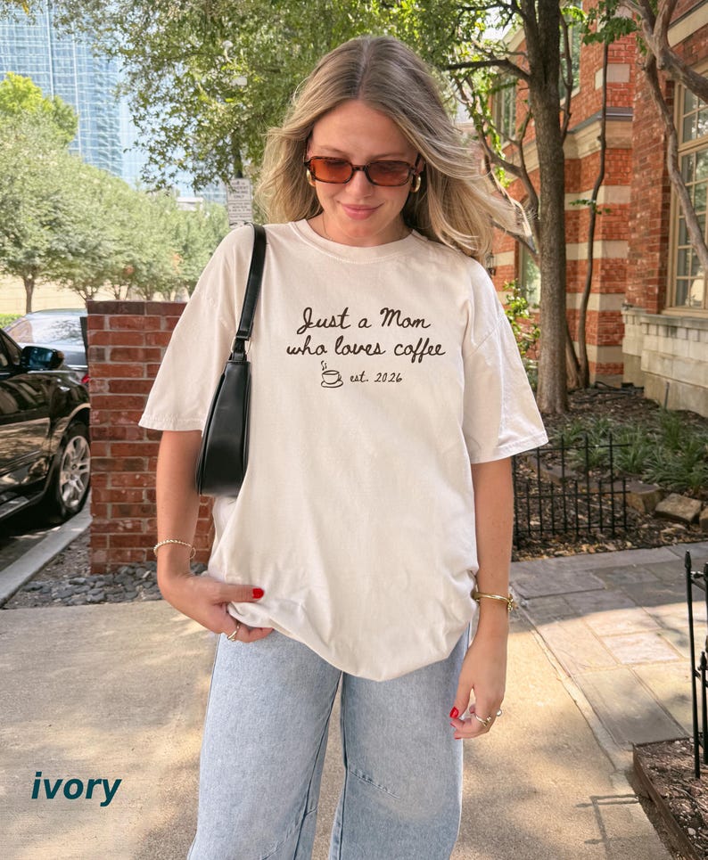 May include: An ivory short-sleeve t-shirt featuring the black text Just a Mom who loves coffee, a small coffee cup icon, and the text est. 2026. The shirt is paired with light blue denim jeans and a black shoulder bag with a thin strap.