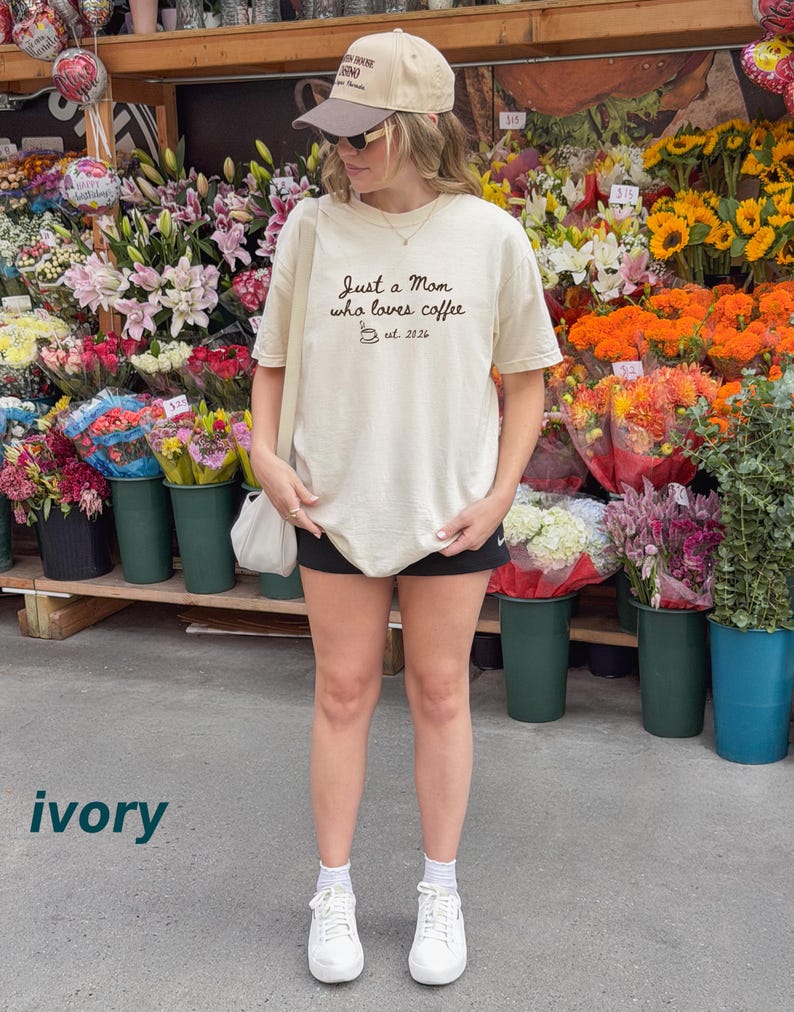 May include: An ivory short-sleeve t-shirt with black text that reads Just a Mom who loves coffee est. 2026, featuring a small coffee cup graphic. The shirt is paired with a beige and brown baseball cap, black shorts, white socks, and white trainers.