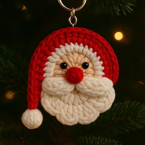 Könnte beinhalten: Handgefertigter Weihnachtsschmuck in Form eines Weihnachtsmann-Gesichts. Der Schmuck ist mit rotem, weißem und beigem Garn gehäkelt und hat eine rote Mütze, einen weißen Bart und eine runde rote Nase. Er hängt an einem goldfarbenen Schlüsselring.