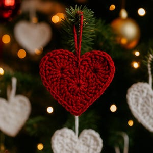 以下が含まれることがあります： クリスマスツリーに吊るされた赤いかぎ針編みのハートのオーナメント。ハートは赤い糸でできており、質感があります。他の白いかぎ針編みのハートのオーナメントと金のオーナメントも表示されます。