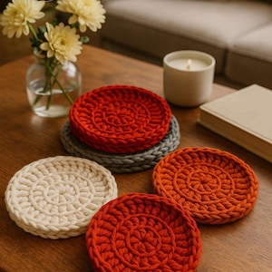 May include: A collection of handmade crochet coasters in various colors, including red, orange, gray, and cream, arranged on a wooden table. The coasters have a circular design with a spiral pattern in the center. The image also includes a vase of yellow flowers, a candle, and a book.