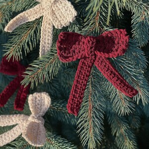 Op de afbeelding: Close-up van een kerstboom met verschillende handgemaakte gehaakte strikversieringen. De strikken zijn in cr&egrave;me- en bordeaux tinten en zijn bevestigd aan de groenblijvende takken. Een feestelijke vakantiedecoratie.