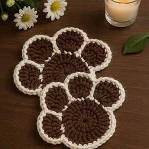 May include: Two brown and white paw-shaped crochet coasters are displayed on a wooden surface. The coasters are surrounded by a lit candle in a glass holder, white daisies, and a green leaf. The coasters are a decorative item.