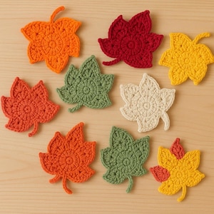 Puede incluir: Una colección de hojas de ganchillo hechas a mano en varios colores otoñales, incluyendo naranja, rojo, verde, amarillo y crema. Las hojas están dispuestas sobre una superficie de madera marrón claro, mostrando sus intrincados detalles de puntada.