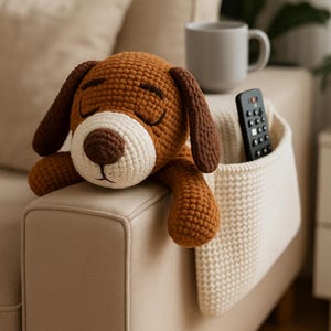 Puede incluir: Un peluche de perro tejido en marrón y blanco con los ojos cerrados descansa sobre un reposabrazos de sofá beige. También se ven un soporte para mando a distancia color crema y una taza. El perro tiene orejas y nariz marrones.