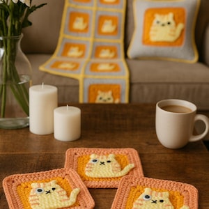 Könnte beinhalten: Gemütliche Szene mit handgefertigten Häkelartikeln. Das Bild zeigt eine Decke, ein Kissen und Untersetzer, alle mit einem gelben Katzendesign auf orangefarbenem und grauem Hintergrund. Weiße Kerzen und eine Kaffeetasse vervollständigen die warme, einladende Ästhetik.