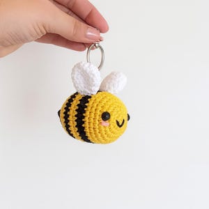 Può includere: Un portachiavi a forma di ape all'uncinetto con corpo giallo, strisce nere e ali bianche. L'ape ha una faccia sorridente con occhi neri e guance rosa. Il portachiavi è tenuto da un anello d'argento.