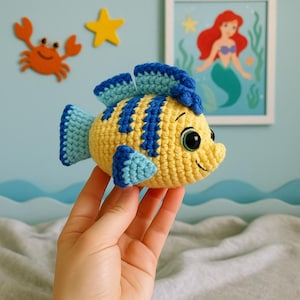 May include: A yellow and blue crocheted fish with large black eyes and a smiling mouth. The fish has blue fins and stripes. The fish is a handmade item, possibly a toy or decorative item.