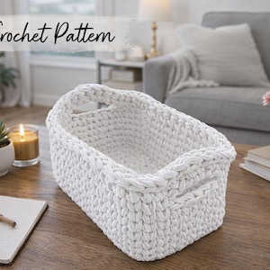 Peut inclure: Un panier rectangulaire blanc en crochet avec des poignées, présenté sur une table en bois. Le panier est fait de fil épais et texturé. Les mots "Crochet Pattern" sont écrits en cursive en haut de l'image. Un cactus, une bougie et un carnet sont également sur la table.