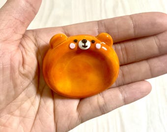 May include: A small, glossy, orange ceramic dish shaped like a bear face. The dish features two rounded ears, a white muzzle with a black nose and mouth, two small black eyes, and two light pink circular cheeks on a smooth, vibrant orange surface.