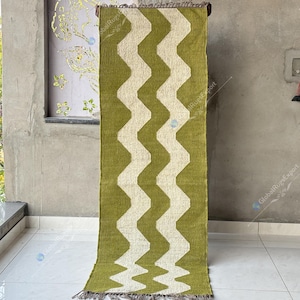 May include: A green and white zigzag patterned rug. The rug is rectangular and has a repeating wave-like design in white against a green background. The rug is hanging on a wooden rod.