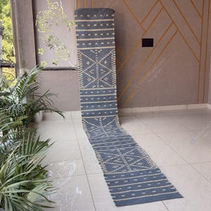 Puede incluir: Una alfombra de pasillo larga y rectangular con una base de color azul pizarra y un patrón geométrico repetitivo en blanco crema. El diseño consta de secciones alternas de intrincados motivos de diamantes y líneas horizontales discontinuas.