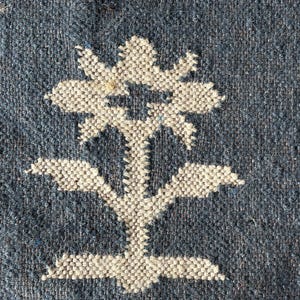 May include: Close-up of a woven textile with a blue background and a large white floral design. The flower has multiple petals and a stem with leaves. The texture of the fabric is visible, showing the weave of the threads.