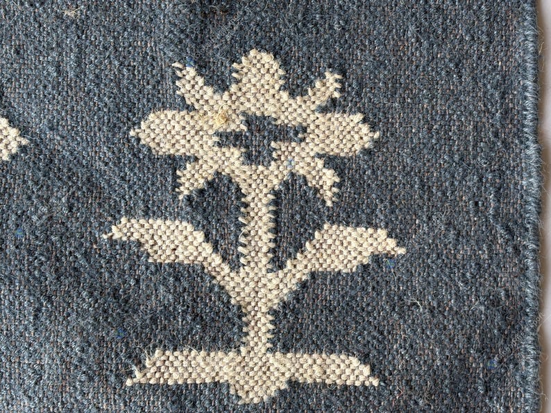 May include: Close-up of a woven textile with a blue background and a large white floral design. The flower has multiple petals and a stem with leaves. The texture of the fabric is visible, showing the weave of the threads.