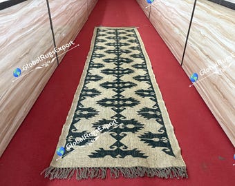May include: A rectangular jute runner rug with a geometric pattern in dark teal and beige. The rug has a fringed edge and is displayed on a red surface. The rug is a decorative home decor item.