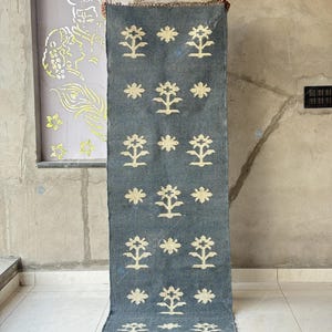 May include: A rectangular, blue-gray rug with a repeating pattern of stylized cream-colored floral motifs. The rug has a fringed edge and is displayed vertically. The background is a neutral-toned wall and floor.