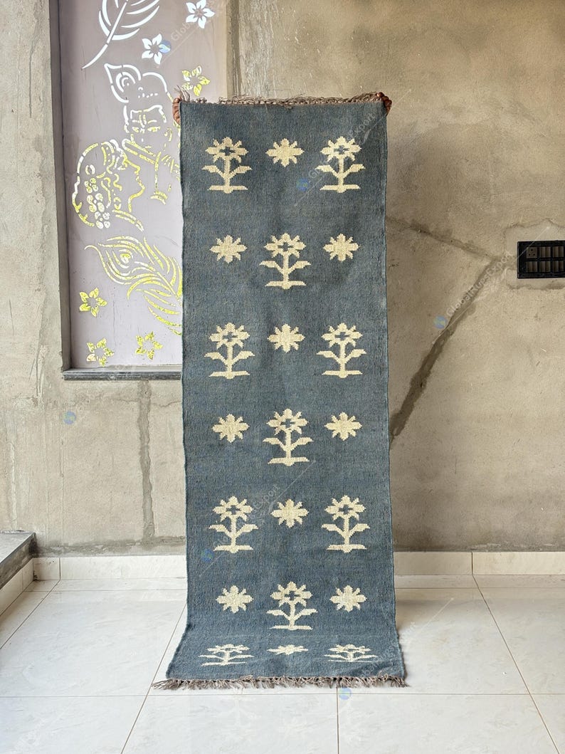May include: A rectangular, blue-gray rug with a repeating pattern of stylized cream-colored floral motifs. The rug has a fringed edge and is displayed vertically. The background is a neutral-toned wall and floor.