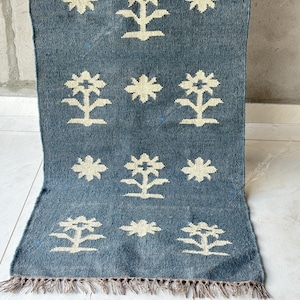 May include: A rectangular, blue rug with a cream-colored floral pattern. The rug has a fringe along the bottom edge. The floral design is repeated across the surface. The rug is made of a woven material.