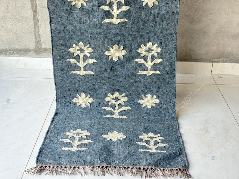 May include: A rectangular, blue rug with a cream-colored floral pattern. The rug has a fringe along the bottom edge. The floral design is repeated across the surface. The rug is made of a woven material.