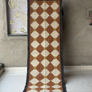 May include: A rectangular brown and cream-colored rug with a geometric diamond pattern. The rug has a dark gray border and fringe along the short ends. The rug is displayed vertically against a neutral-colored wall.