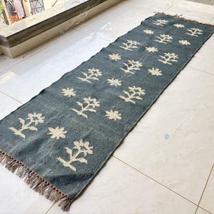 May include: A rectangular, blue runner rug with a repeating pattern of cream-colored floral and star-like designs. The rug has a fringed edge and is displayed on a tiled floor. The rug is approximately 60 inches long.