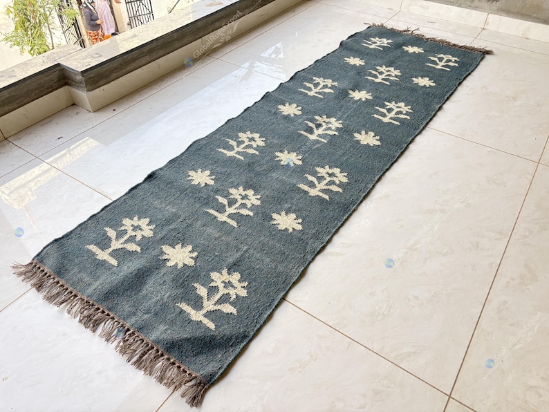 May include: A rectangular, blue runner rug with a repeating pattern of cream-colored floral and star-like designs. The rug has a fringed edge and is displayed on a tiled floor. The rug is approximately 60 inches long.
