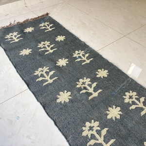 May include: A rectangular, dark gray rug with a repeating pattern of cream-colored floral and tree-like designs. The rug has a fringed edge and is placed on a light-colored tiled floor. The rug is a decorative home accent.