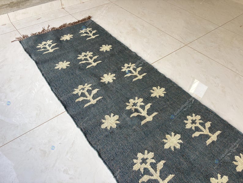 May include: A rectangular, dark gray rug with a repeating pattern of cream-colored floral and tree-like designs. The rug has a fringed edge and is placed on a light-colored tiled floor. The rug is a decorative home accent.