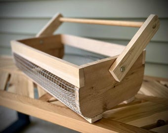 May include: A light-colored wooden basket with a wire mesh front and a wooden handle. The basket has a rectangular shape with rounded edges and is constructed with visible screws and bolts. The handle is attached to the sides with angled wooden supports.