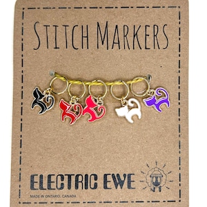 Könnte beinhalten: Eine Reihe von Maschenmarkierern auf einer braunen Karte. Die Marker sind goldfarbene Ringe mit farbigen Anhängern in Schwarz, Rot, Weiß und Lila. Auf der Karte stehen die Wörter "STITCH MARKERS" und "ELECTRIC EWE" in schwarzer Schrift.