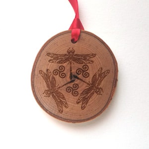 Könnte beinhalten: Runder Holzschmuck mit rotem Band zum Aufhängen. Er zeigt drei gravierte Libellen und ein zentrales Design. Das Holz hat eine natürliche Maserung und ist hellbraun.