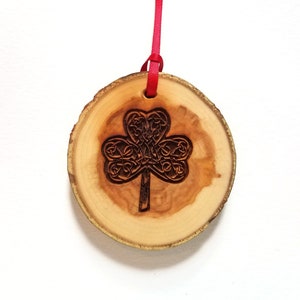 May include: A wooden ornament shaped like a circle with a Celtic knot shamrock design carved into the surface. The ornament is attached to a red ribbon.