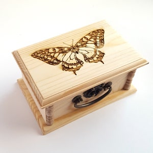 Puede incluir: Una pequeña caja de madera con un diseño de mariposa quemado en la tapa. La caja tiene una manija de metal y adornos decorativos alrededor de los bordes.