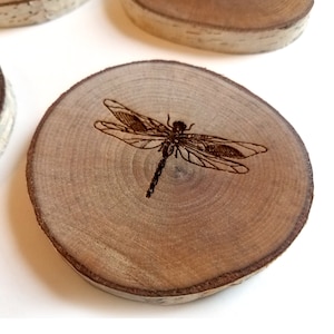 Może przedstawiać: Drewniana podkładka pod kubek z laserowo grawerowanym wzorem ważki. Podkładka jest okrągła i ma naturalne usłojenie drewna.