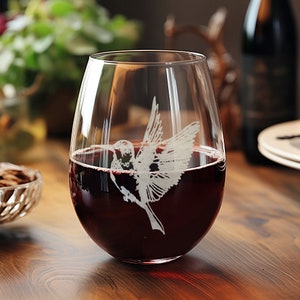 May include: A clear glass wine glass with a hummingbird etched into the side. The glass is filled with red wine.