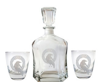 May include: A clear glass decanter with a stopper and two matching glasses. Each piece features an etched design of a raven inside a Celtic knot.
