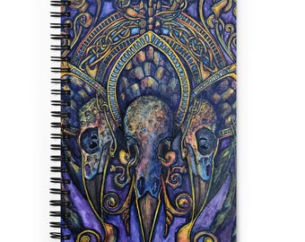 Puede incluir: Un cuaderno de espiral con una ilustración colorida de tres cráneos de pájaros rodeados de intrincados diseños dorados y morados.