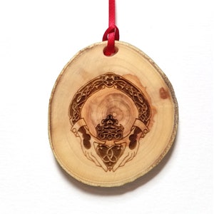 Op de afbeelding: Een houten ornament met een Keltisch knoopontwerp, een kroon, handen en een hart. Het ornament is bevestigd aan een rood lint.